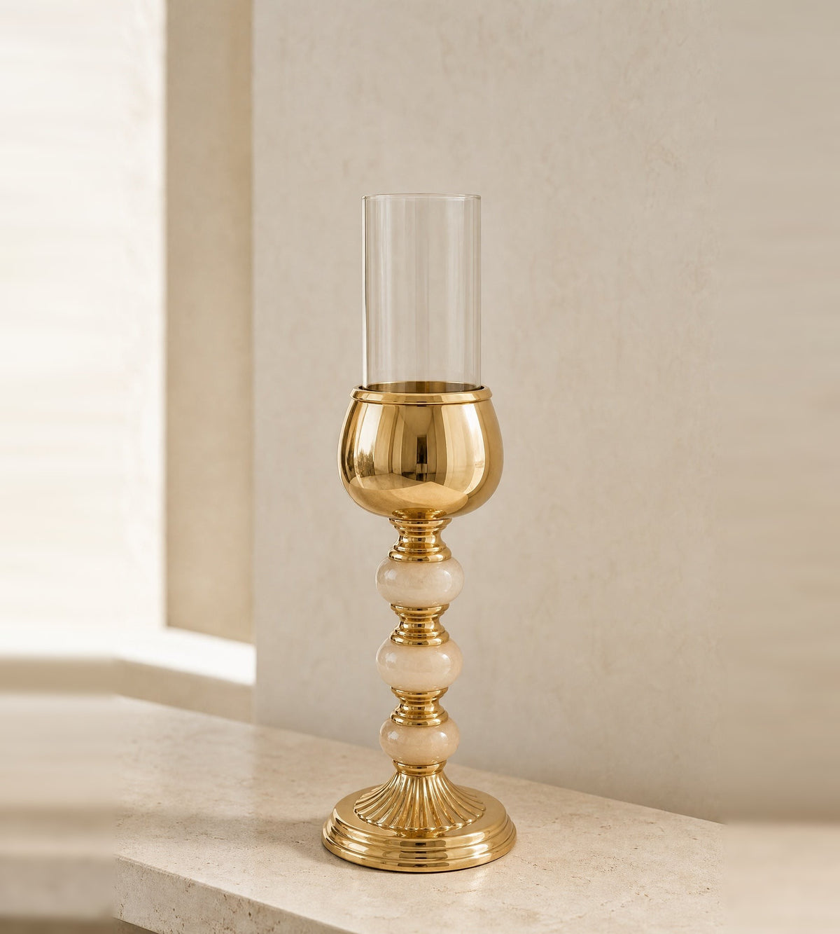 Gold-finish hurricane candle holder with three onyx spheres in the stem and a clear glass shade, on a travertine console