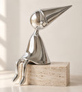 Silver chrome shelf-sitter sculpture with a pointed cone hat, legs hanging over a travertine block