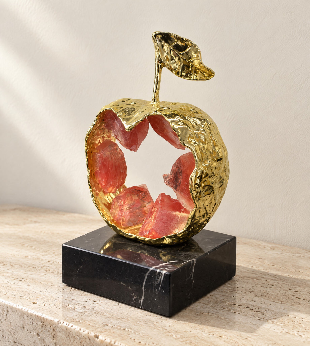 Gold metal apple sculpture with an open hammered shell holding red K9 crystal, on a black marble base, on travertine
