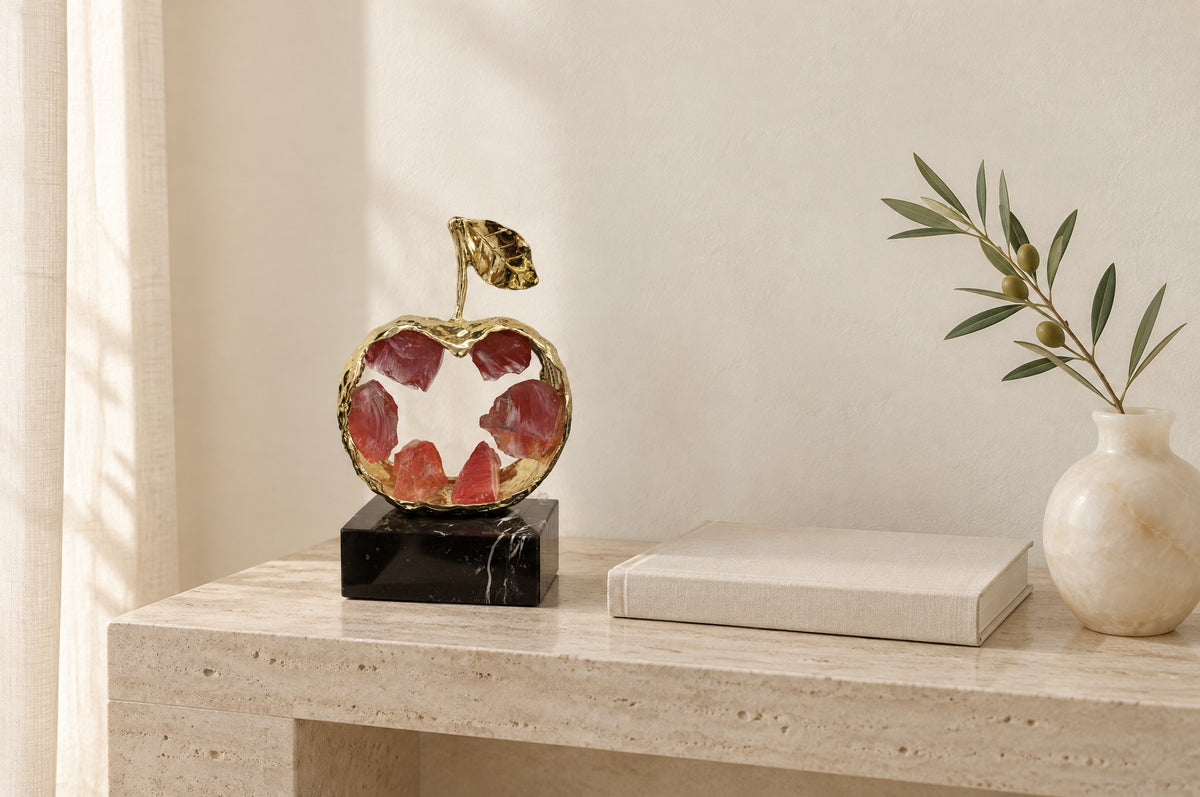Gold metal apple sculpture with red K9 crystal on a travertine console beside a linen book and an olive stem, showing its 23 cm size