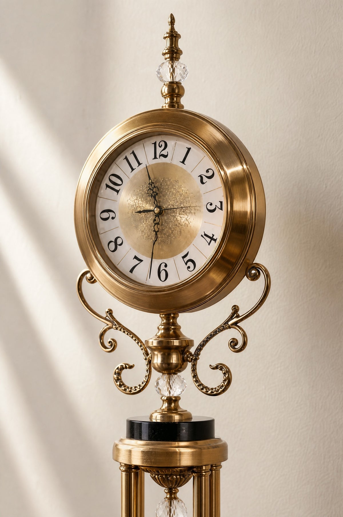 Close view of the clock head: ivory dial with black Arabic numerals behind glass, brass bezel, crystal-set finial and scrolled brass arms