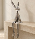 Silver chrome resin bunny sculpture sitting on the edge of a pale travertine console, long legs hanging down