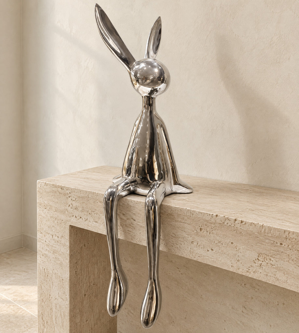 Silver chrome resin bunny sculpture sitting on the edge of a pale travertine console, long legs hanging down