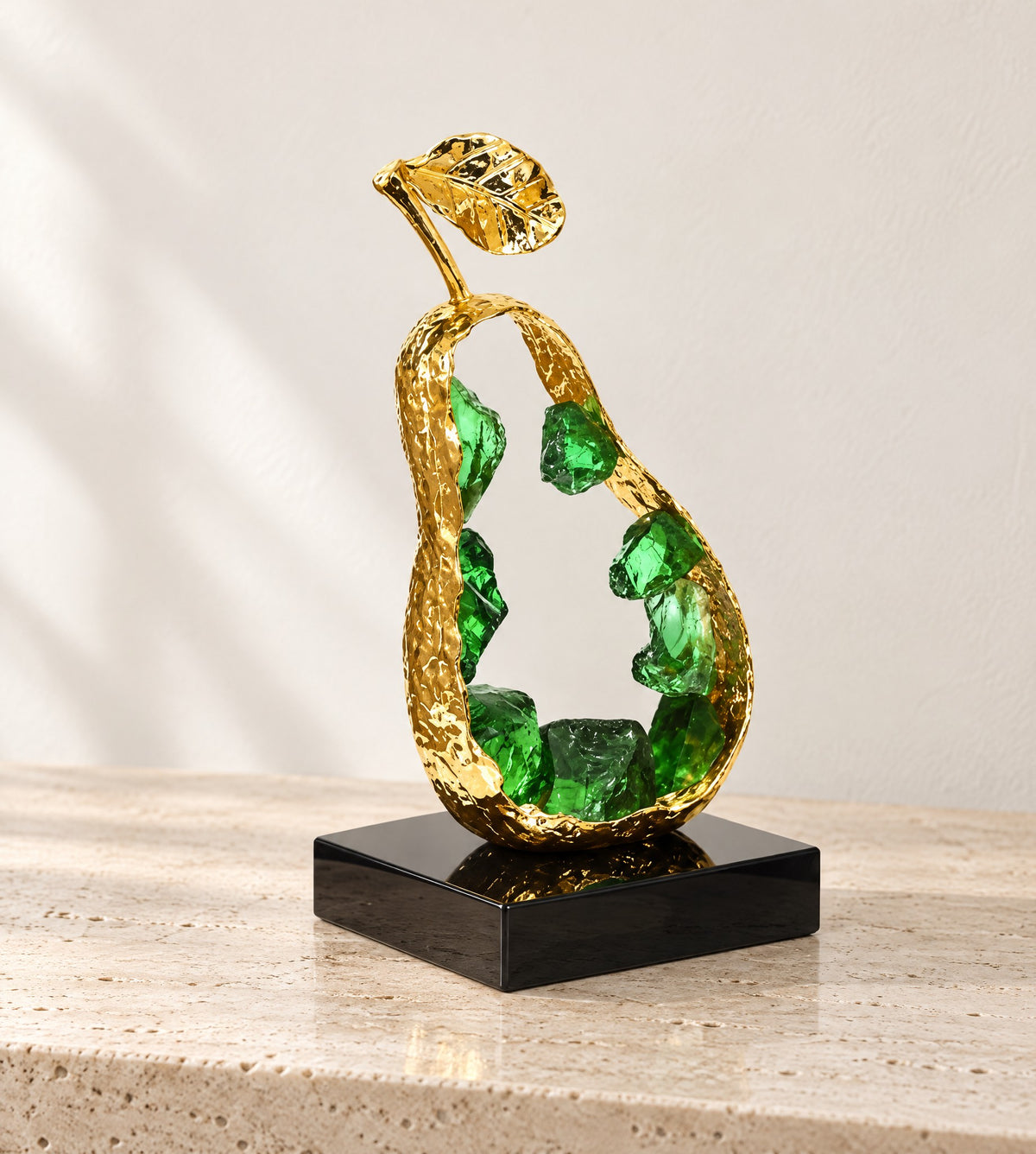 Gold metal pear sculpture with an open hammered shell holding green K9 crystal, on a black base, on travertine