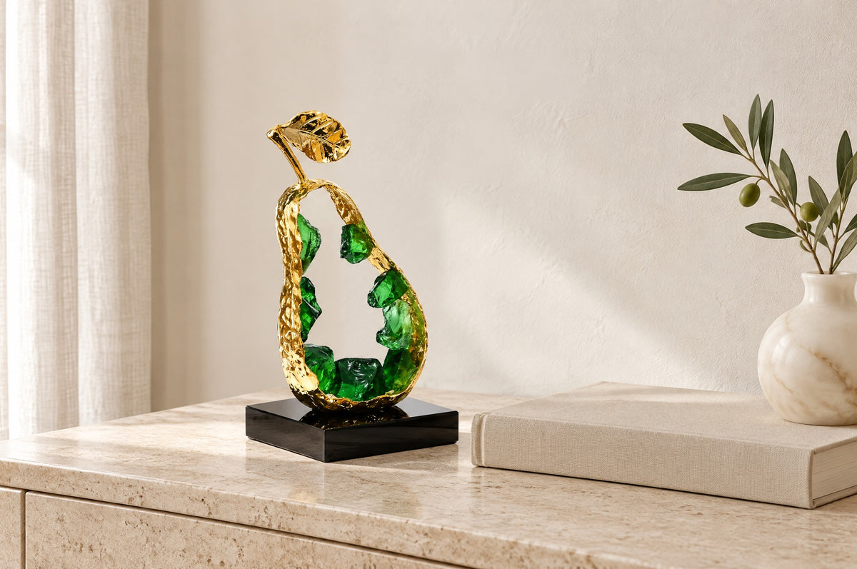 Gold pear sculpture with green K9 crystal on a travertine console beside a linen book and an olive stem, showing its 25 cm size